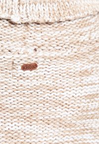Dzianina Khujo w beżowo-kremowej odsłonie z teksturowanym wzorem. Zawiera małą brązową skórzaną metkę z logo marki.