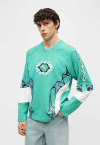 Νεαρός με σγουρά μαλλιά φοράει μια μπλούζα μακρυμάνικη σε τυρκουάζ χρώμα με αφηρημένα μαύρα και λευκά σχέδια και λογότυπο Puma, συνδυασμένη με ανοιχτόχρωμο μπλε τζιν.