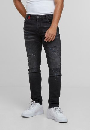 Mann trägt schwarze, abgenutzte Jeans mit Farbspritzern und weiße Sneakers, steht vor einem grauen Hintergrund.