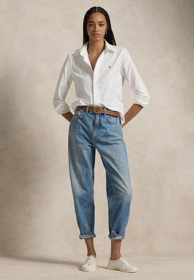 Ung kvinne står med hendene bak ryggen, iført hvit skjorte med knapper, lyseblå oppbrettede jeans, brunt belte og hvite joggesko.