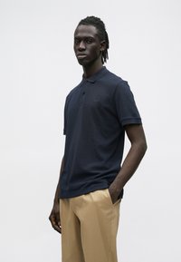 Egy dreadlock hajú férfi, sötétkék pólóinget és bézs nadrágot visel, egyik kezét a zsebében tartva áll egy egyszínű, világos háttér előtt.