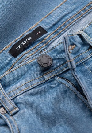 Jeans in denim di un blu chiaro con cuciture gialle a contrasto. Presentano un'etichetta nera, chiusura con zip e un bottone nero rotondo.