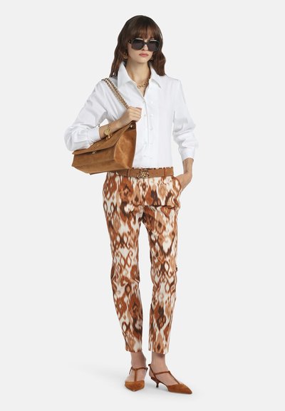 Femme portant de grandes lunettes de soleil noires, une chemise blanche boutonnée, un pantalon marron à motifs, des talons beiges, et portant un sac à main en daim beige avec une bandoulière chaîne dorée.
