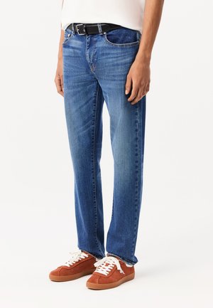 Blauwe denim jeans met een rechte pasvorm, met subtiele vervaging, vijf zakken en een zwarte riem. Gecombineerd met oranje suède sneakers.