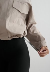 Hand past het trekkoord van een beige jas bij de taille aan over zwarte geribbelde leggings en benadrukt de zak- en manchetdetails van de jas.