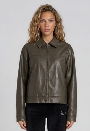 Femme aux cheveux blonds ondulés portant une veste en cuir marron foncé avec fermeture éclair et un pantalon noir, debout devant un fond clair uni.