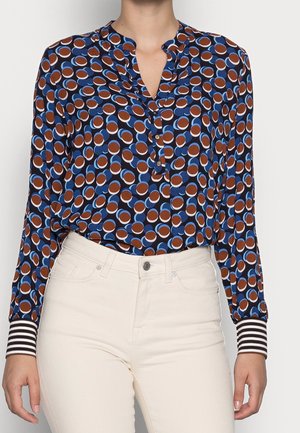 Femme portant une blouse boutonnée avec des motifs circulaires marron, blanc et bleu, des poignets rayés, rentrée dans un pantalon taille haute écru.