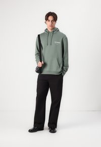 Hooded, green sweatshirt with front pocket and white logo; paired with black pants and a dark crossbody bag; sleek, minimal design.