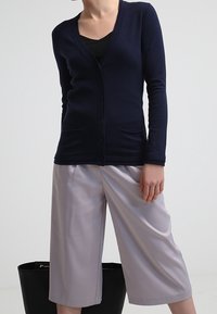 Cardigan blu navy con bottoni e tasche, abbinato a pantaloni a gamba larga grigio chiaro, che creano una silhouette semplice e pulita.