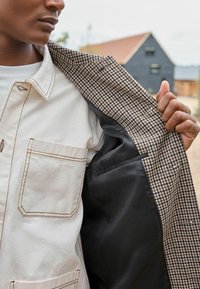 Beige takki, jossa on houndstooth-kuvioinen vuori. Sisätasku, joka on valmistettu sileästä mustasta kankaasta. Raskas rakenne ja jämäkkä muotoilu.
