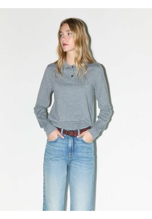 Jonge vrouw die een grijze polo met lange mouwen en lichtblauwe spijkerbroek met een bruine riem draagt, staat met haar handen in de zakken en kijkt opzij.