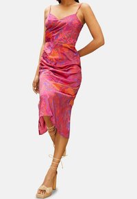 Nainen, joka pukeutunut vaaleanpunaiseen ja oranssiin kukalliseen hihattomaan midimekkoon, jossa on rypytyksiä, ja beigeihin nilkkaremmeill ä varustettuihin korkokenkiin.