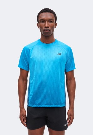 Homme debout face à l'avant portant un t-shirt de sport bleu vif à manches courtes et un short de sport noir sur un fond uni.