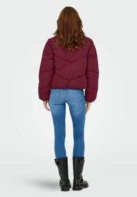 Burgundska pufasta jakna s chevron kvadratnim uzorkom, skraćeni dizajn, u kombinaciji s plavim uskim trapericama i crnim čizmama do koljena s kopčama.