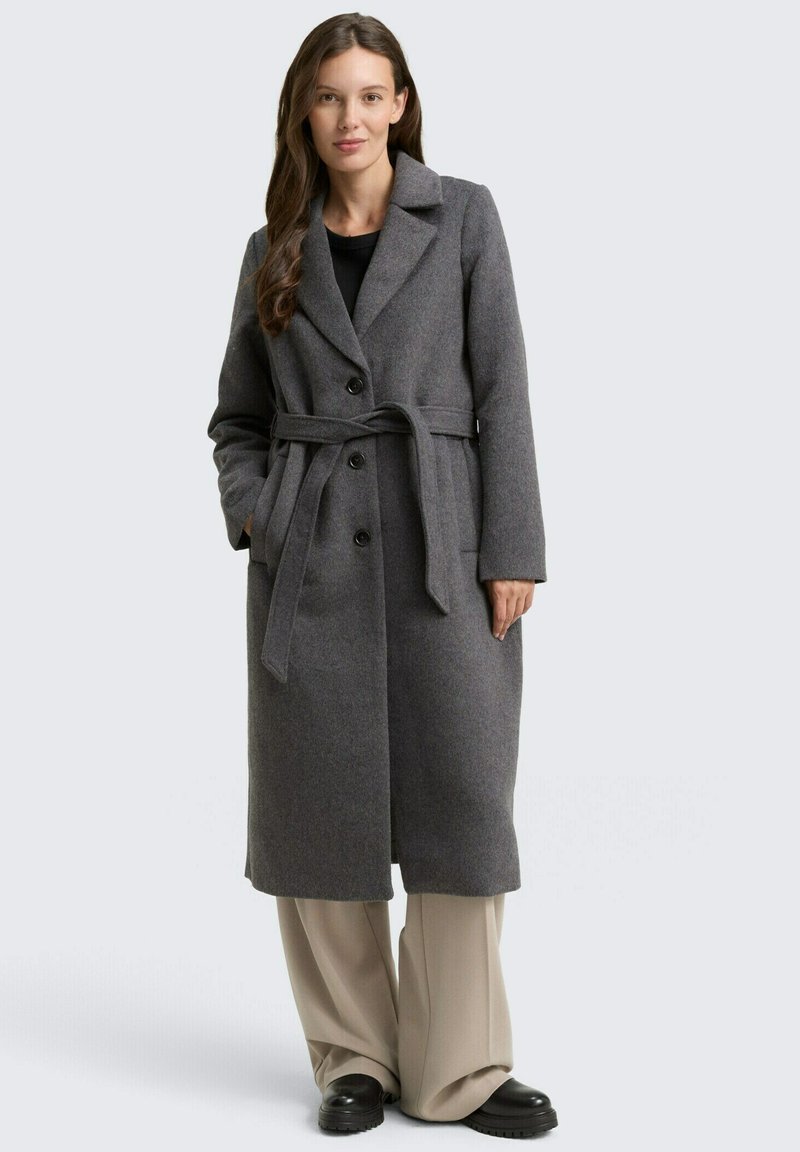 Vrouw in een lange grijze jas met riem over een zwart top, beige wijde broek, en zwarte schoenen, staand met handen in de zakken.