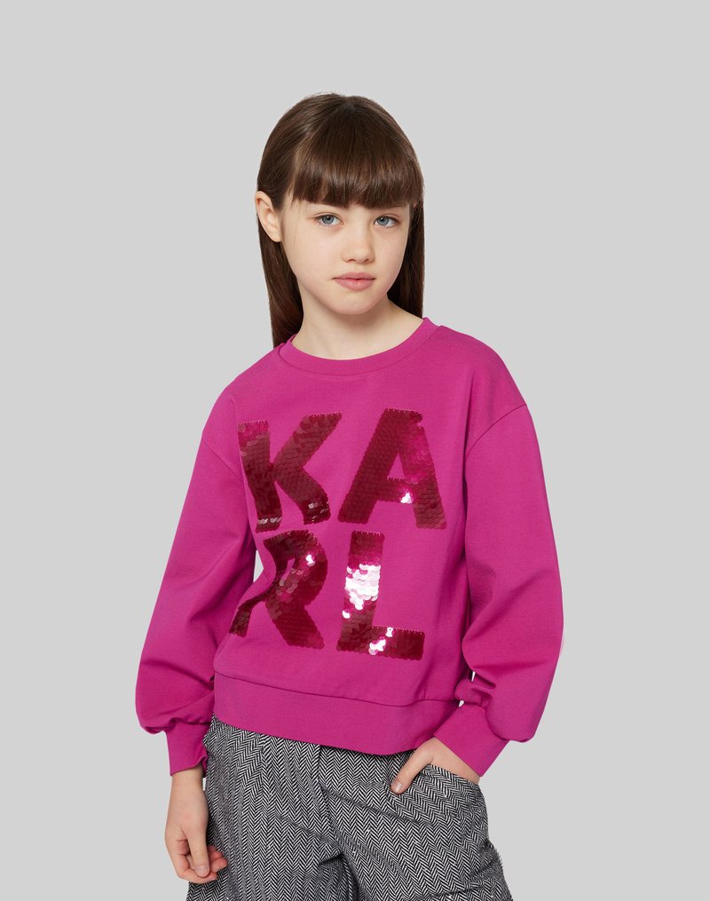 Fuksjowa bluza z napisem "KARL" wyszywanym cekinami. Ma okrągły dekolt i bufiaste rękawy. Zestawiona z spodniami w wzór houndstooth.