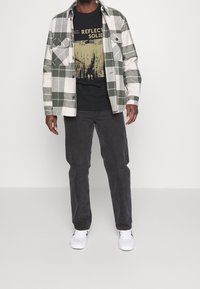 Randig grön och vit flanellskjorta över en svart t-shirt med tryck, kombinerad med mörka jeans och vita sneakers.