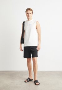 Camisa blanca sin mangas con el texto "Esto es Amor", pantalones cortos negros con logo, sandalias negras. Diseño minimalista y ajuste relajado.