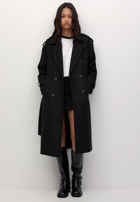 Žena stoji, odjevena u dugi crni dvostruko zakopčani kaput, bijelu košulju, crne šortse i crne čizme do koljena, nasuprot jednostavne pozadine.