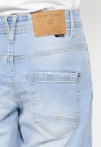 Jean en denim bleu clair montrant la poche arrière et un patch en cuir marron avec "Koroshi Brand" et le logo au-dessus de la poche.