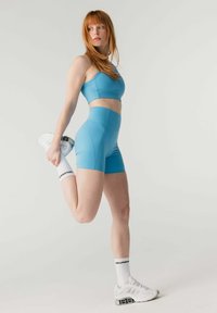 Sinistes spordiriietes naine venitab reielihast, hoides kannast käe taga, jalas valged tossud ja "GIRLFRIEND" sokid, hele taust.