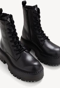 Bottines en cuir noir avec un laçage sur le devant, une fermeture éclair sur le côté, une semelle en caoutchouc épaisse et un détail de couture texturé.