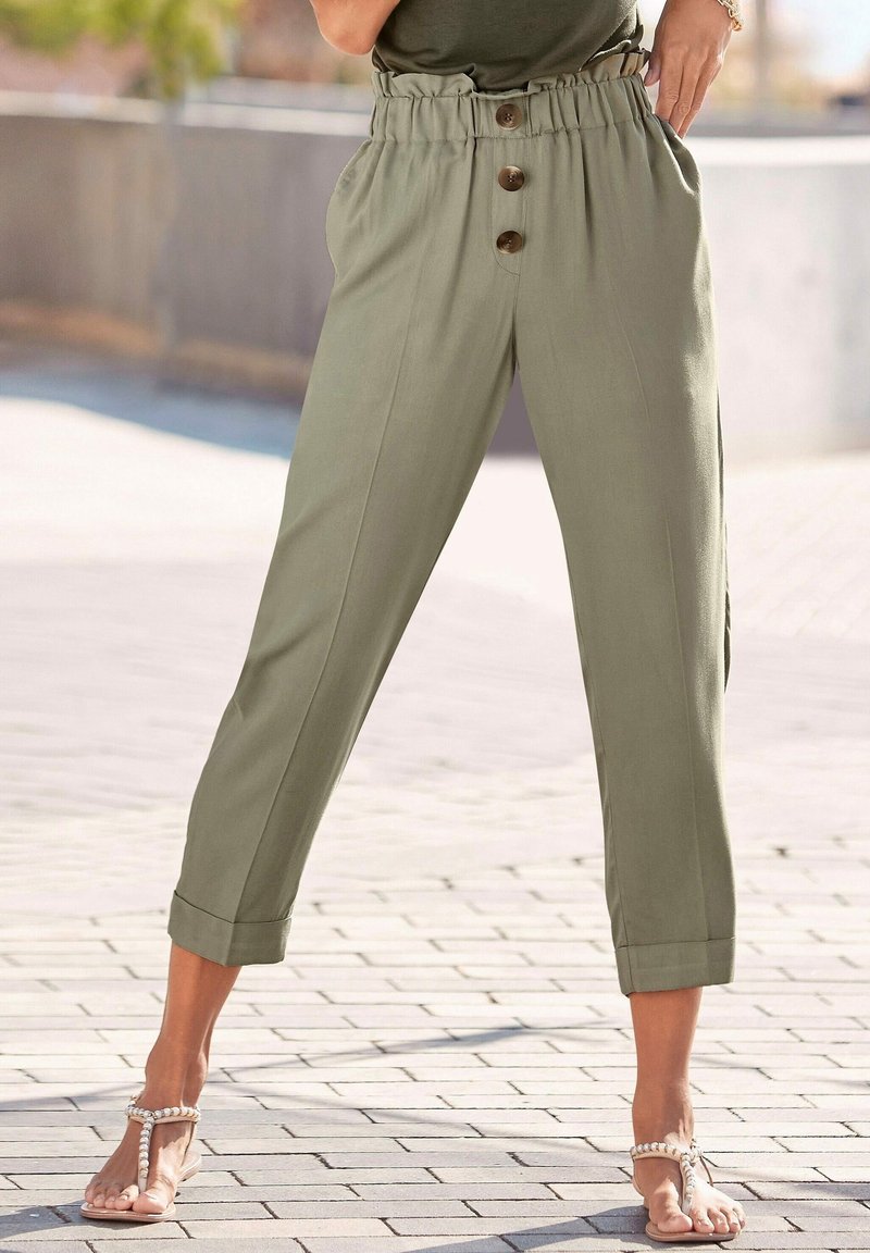 Femme portant un pantalon taille haute vert olive à trois boutons et revers roulés, associé à des sandales ornées de perles dans une rue pavée.
