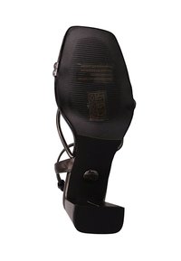 Musta korkokenkä, jossa on ristiin menevät hihnat, teksturoitu pohja ja pyöreä logokuviointi kannassa. Sileä nahkapinta.