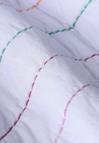 Witte gequiltte stof met kleurrijke stiksels in oranje, groen, roze en blauw, met een zachte, gladde textuur en verhoogd patroon.