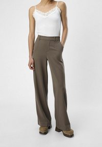 Bílá kamizola s krajkovým lemem v kombinaci s hnědými kalhotami s vysokým pasem a širokými nohavicemi. Kalhoty mají přední kapsy a hladký povrch.