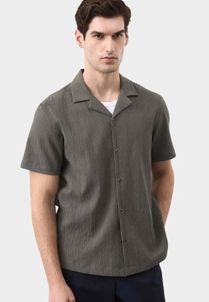 Mężczyzna w teksturowanej oliwkowozielonej koszuli na guziki z krótkim rękawem, na białej podkoszulce, na jasnym, jednolitym tle.