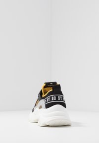 Chaussure de sport noire et jaune avec une tige en tissu texturé, une semelle blanche épaisse et une sangle noire portant l'inscription "90 NY" en blanc.