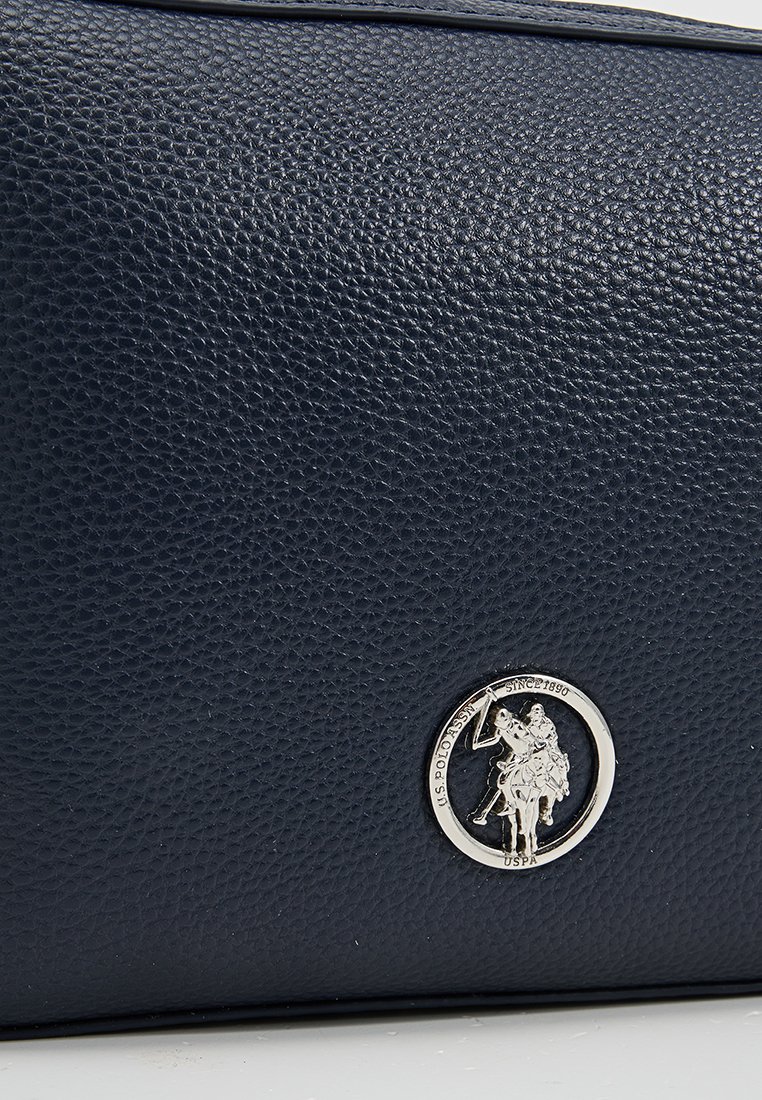 Granatowa torba ze skórzanego materiału o fakturze, z okrągłym, srebrnym logo przedstawiającym gracza polo. Solidna konstrukcja o gładkim wykończeniu.