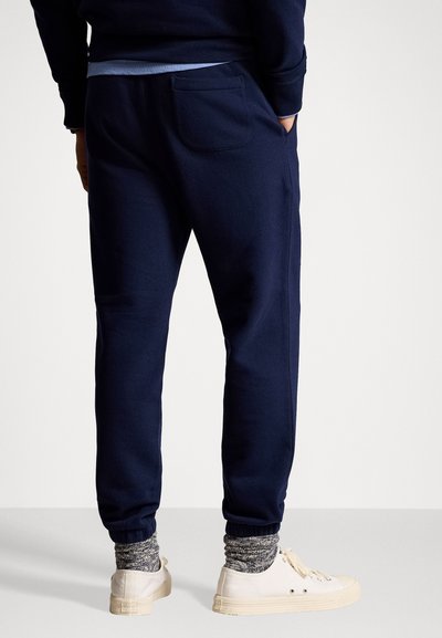 Pantalons de survêtement bleu marine avec des poignets élastiques et une poche arrière, fabriqués dans un matériau doux, associés à des chaussures blanches et des chaussettes grises texturées.