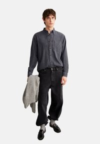 Dunkelgraues Denim-Hemd mit Knöpfen und Brusttasche, kombiniert mit schwarzen lässigen Jeans. Das Model hält einen strukturierten hellgrauen Strickpullover.