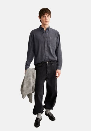 Dunkelgraues Denim-Hemd mit Knöpfen und Brusttasche, kombiniert mit schwarzen lässigen Jeans. Das Model hält einen strukturierten hellgrauen Strickpullover.
