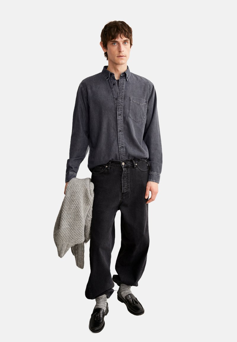 Dunkelgraues Denim-Hemd mit Knöpfen und Brusttasche, kombiniert mit schwarzen lässigen Jeans. Das Model hält einen strukturierten hellgrauen Strickpullover.