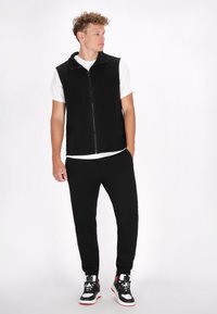 Schwarze ärmellose Weste über einem weißen T-Shirt, kombiniert mit schwarzen Hosen und schwarzen Sneakers mit roten Akzenten. Glattes Material, lässige Passform.