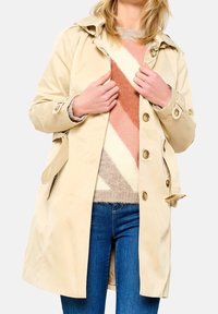 Béžový nepromokavý trenčkot s kapucí, velkými knoflíky a bočními kapsami, vrstvený přes pletený svetr s geometrickými vzory.