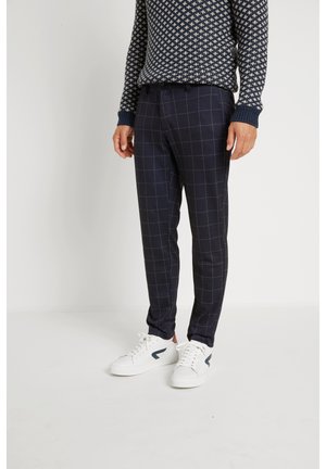 Mężczyzna w granatowych spodniach w kratę windowpane, wzorzystym granatowo-białym swetrze oraz białych sneakersach z czarnymi akcentami.