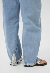 Svetlo modré denimové džínsy s roztrhnutými okrajmi, s voľným strihom a poškodenými miestami na manžetách, doplnené čiernymi šľapkami.