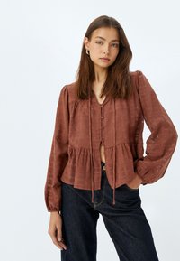 Blusa marrone con texture, scollatura a V, chiusura a bottoni e orlo a peplum. Maniche lunghe con polsini arricciati, abbinata a jeans scuri.