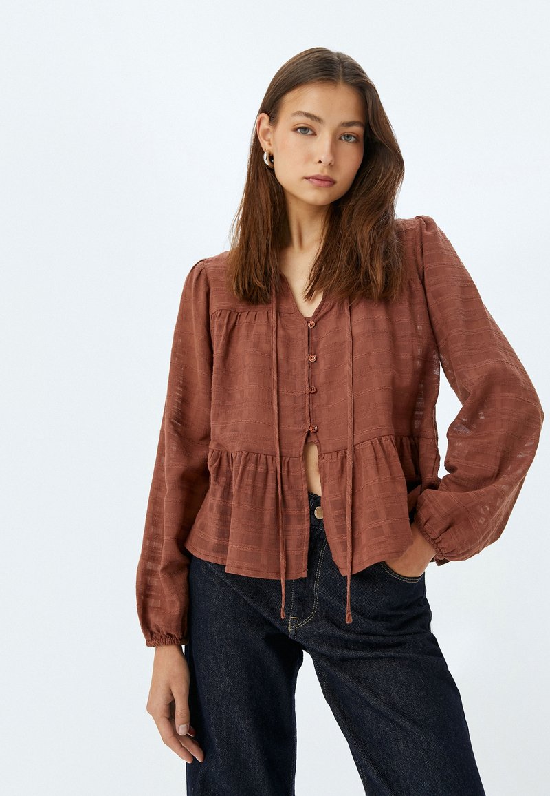 Blusa marrone con texture, scollatura a V, chiusura a bottoni e orlo a peplum. Maniche lunghe con polsini arricciati, abbinata a jeans scuri.