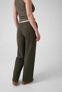 Personne portant un pantalon large olive foncé, des chaussures marron et un haut sans manches olive, debout devant un fond clair uni.