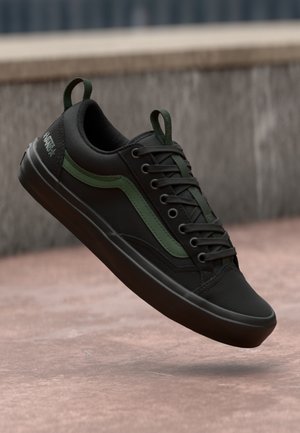 Sort Vans sneaker med grøn sidestribe og trækketapper, der svæver over en lyserød betongrund nær en stenmur.