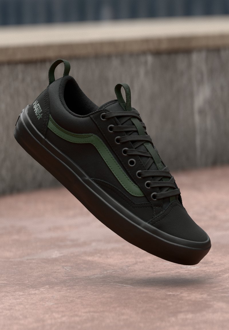 Baskets Vans noires avec bande latérale verte et languettes flottant au-dessus d'une surface en béton rose près d'un mur en pierre.