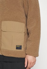 Pullover in pile marrone con una tasca rettangolare beige. La texture è morbida e pelosa, con un piccolo etichetta nera sulla tasca.