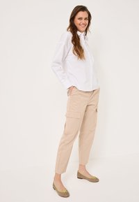 White button-up shirt, beige cargo pants with side pockets, and brown flats with a green accent ribbon. Model stands against a plain background.