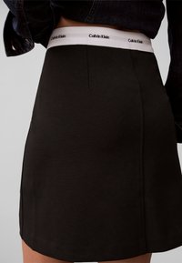 Μαύρη φούστα φορεμένη με λευκή ζώνη Calvin Klein, ορατή κάτω από μια κοντή μαύρη ζακέτα σε ανοιχτό φόντο.