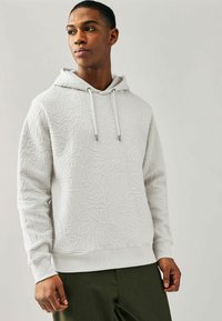 Sudadera blanca con patrón texturizado, capucha con cordones, puños y dobladillo de canalé. Combinada con pantalones verde oliva. Fondo claro.